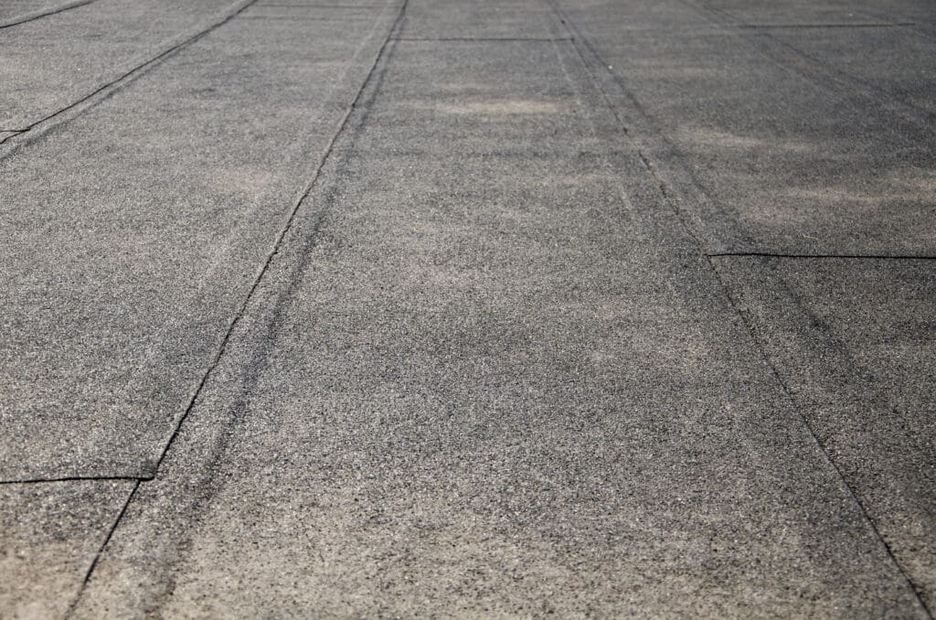 Close-up view of a flat, grey, asphalt or tar roof with visible seams and texture, showing slightly worn and weathered surface.