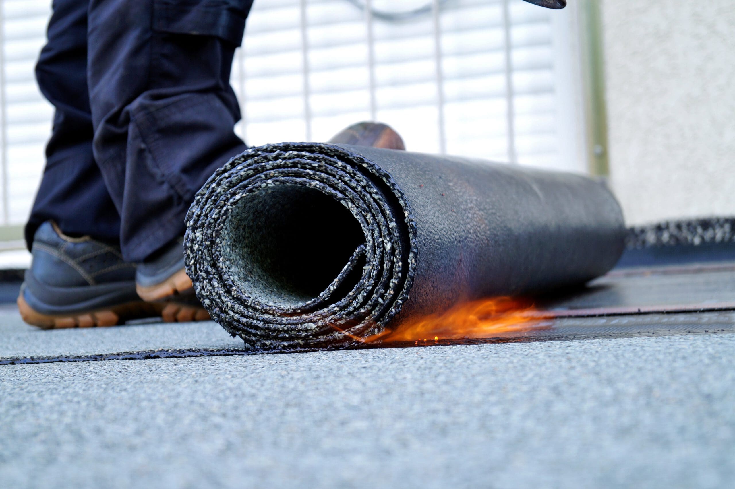 A close-up of a worker's feet next to a rolled roofing material being installed with a flame, melting the underside for adhesion on a flat rooftop.