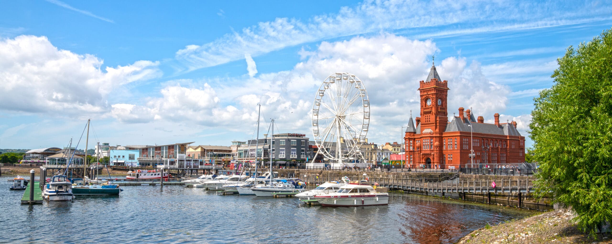 Boats docked in a marina beside a large Ferris wheel and a historic red brick building, with blue sky and scattered clouds overhead. Trees are visible on the right side of the image.