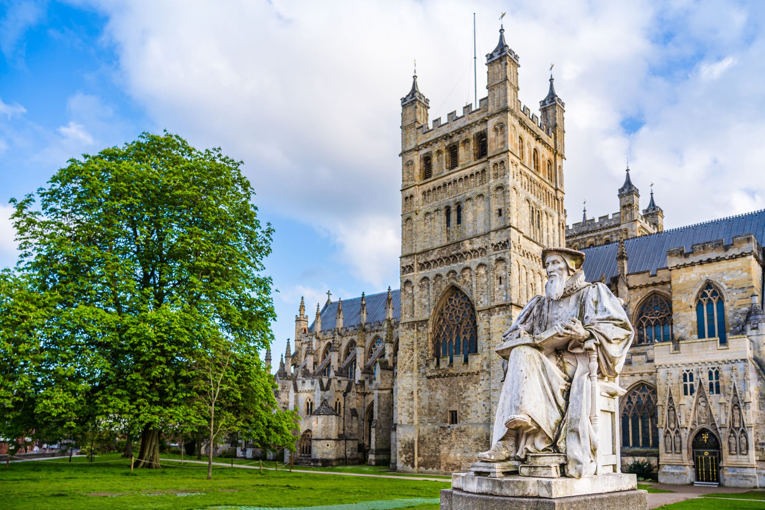 A large stone cathedral with tall towers and arched windows stands beside a leafy green tree and a seated statue of a man in robes, under a partly cloudy sky.