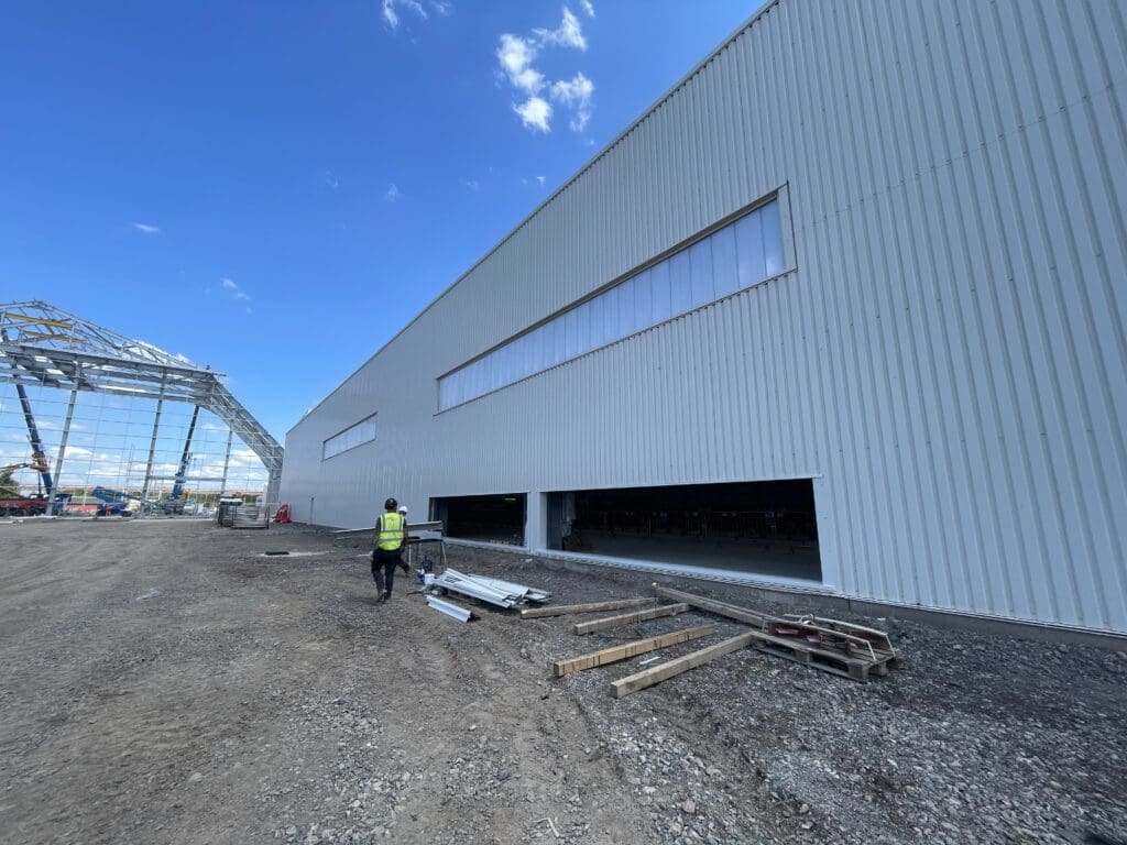 A person in a yellow safety vest walks next to a large industrial building under construction, with piles of materials on the ground and a steel framework visible in the background under a blue sky.