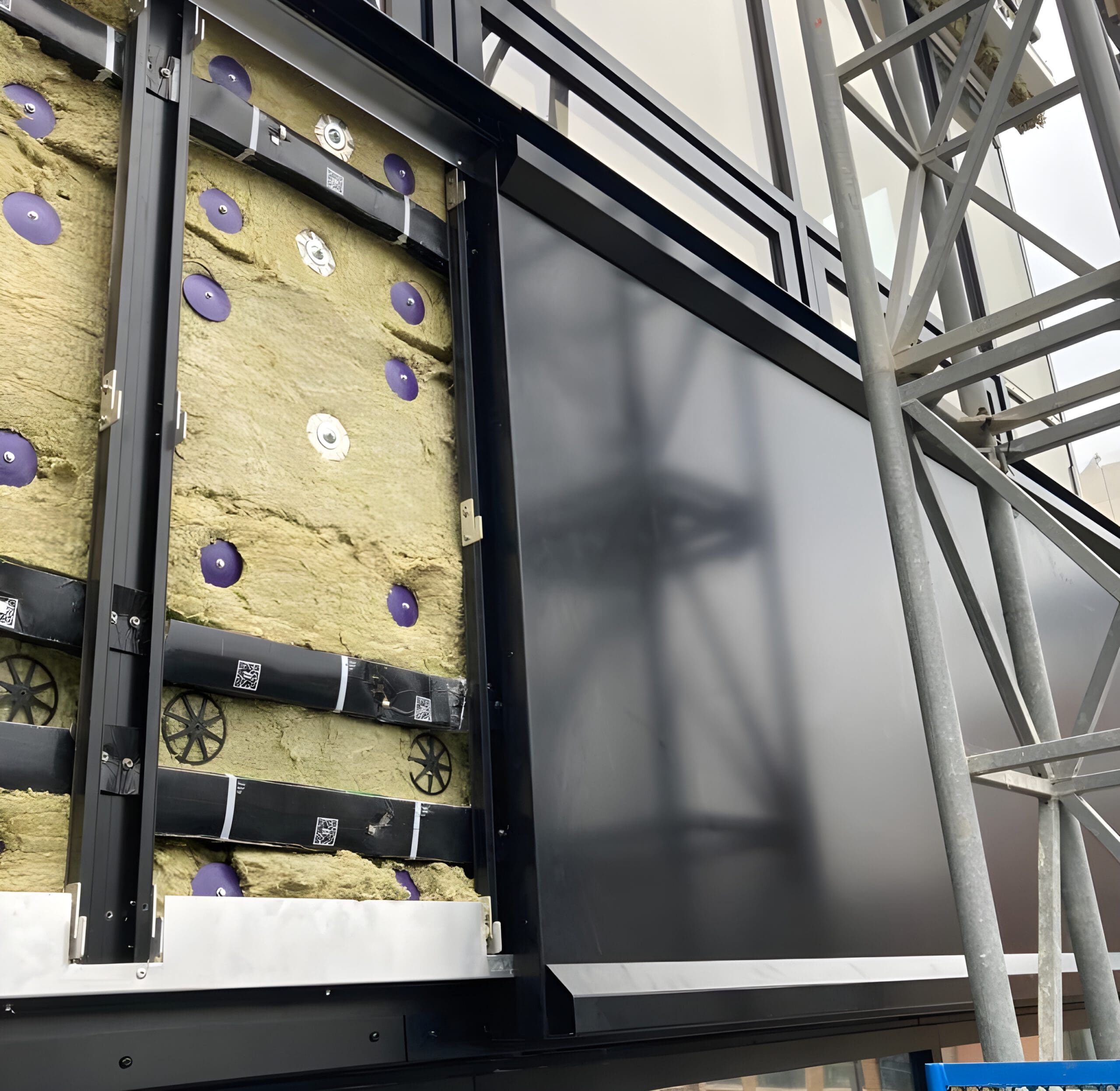 A building facade under construction shows exposed insulation panels on the left side and a smooth, dark metal panel on the right, with scaffolding visible to the right of the image.