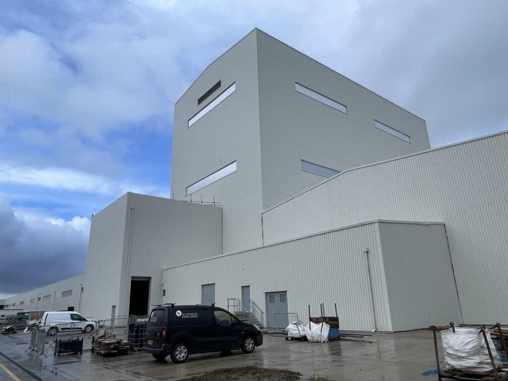 A large, modern white industrial building with tall, rectangular sections and narrow windows. Several vehicles and construction materials are visible on the wet pavement in front. The sky is partly cloudy.