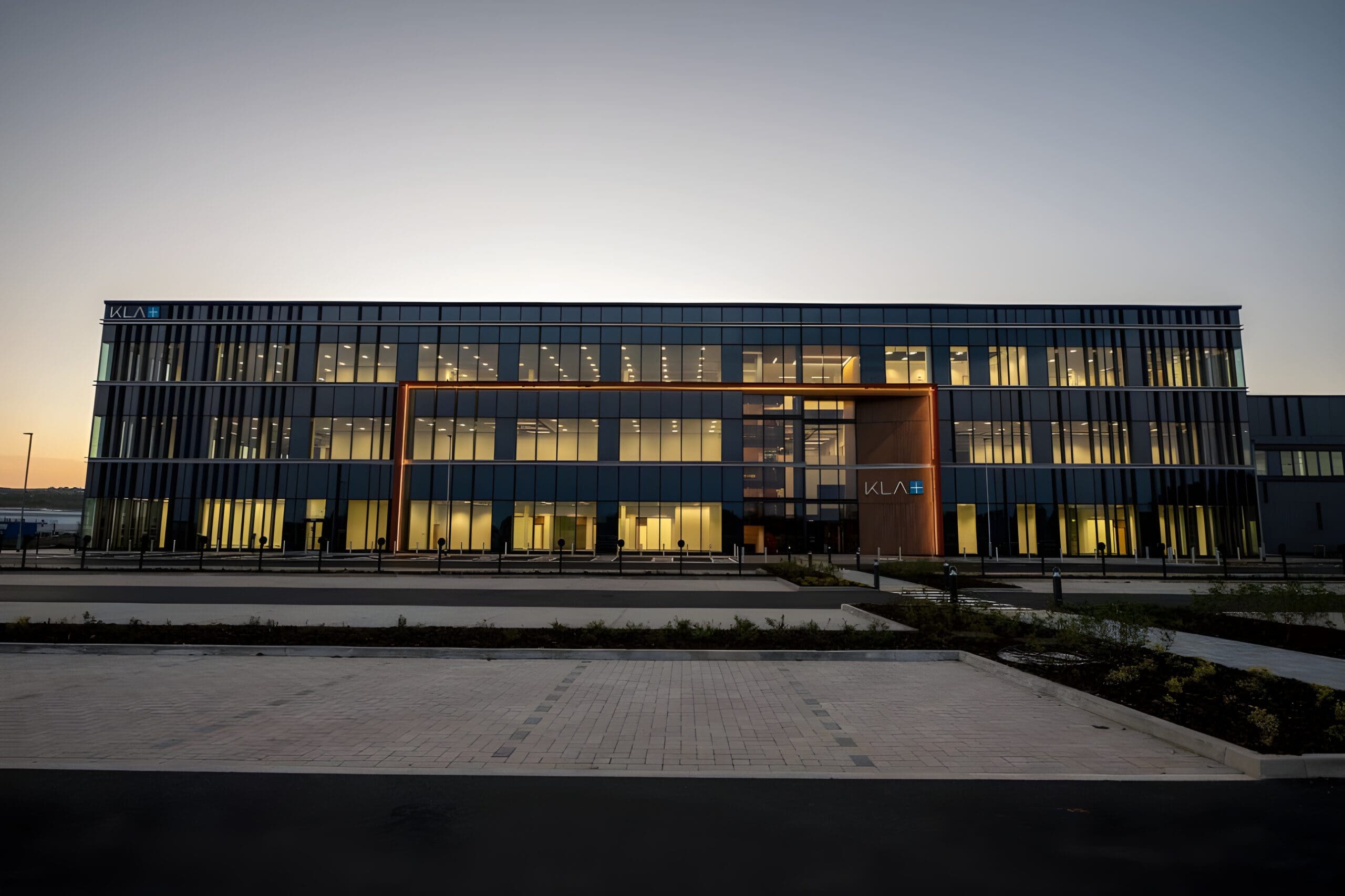 A modern, multi-story office building with large glass windows is illuminated from within at dusk. The building has a sign that reads "KLA" above the entrance, and the area in front is landscaped with walkways.