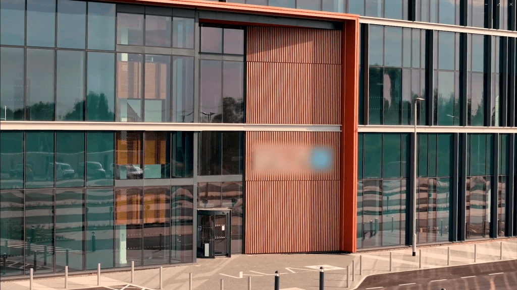 A modern glass office building with large windows and vertical wooden panels. The entrance is visible at the bottom, and a row of bike racks and parking spaces are in front of the building.