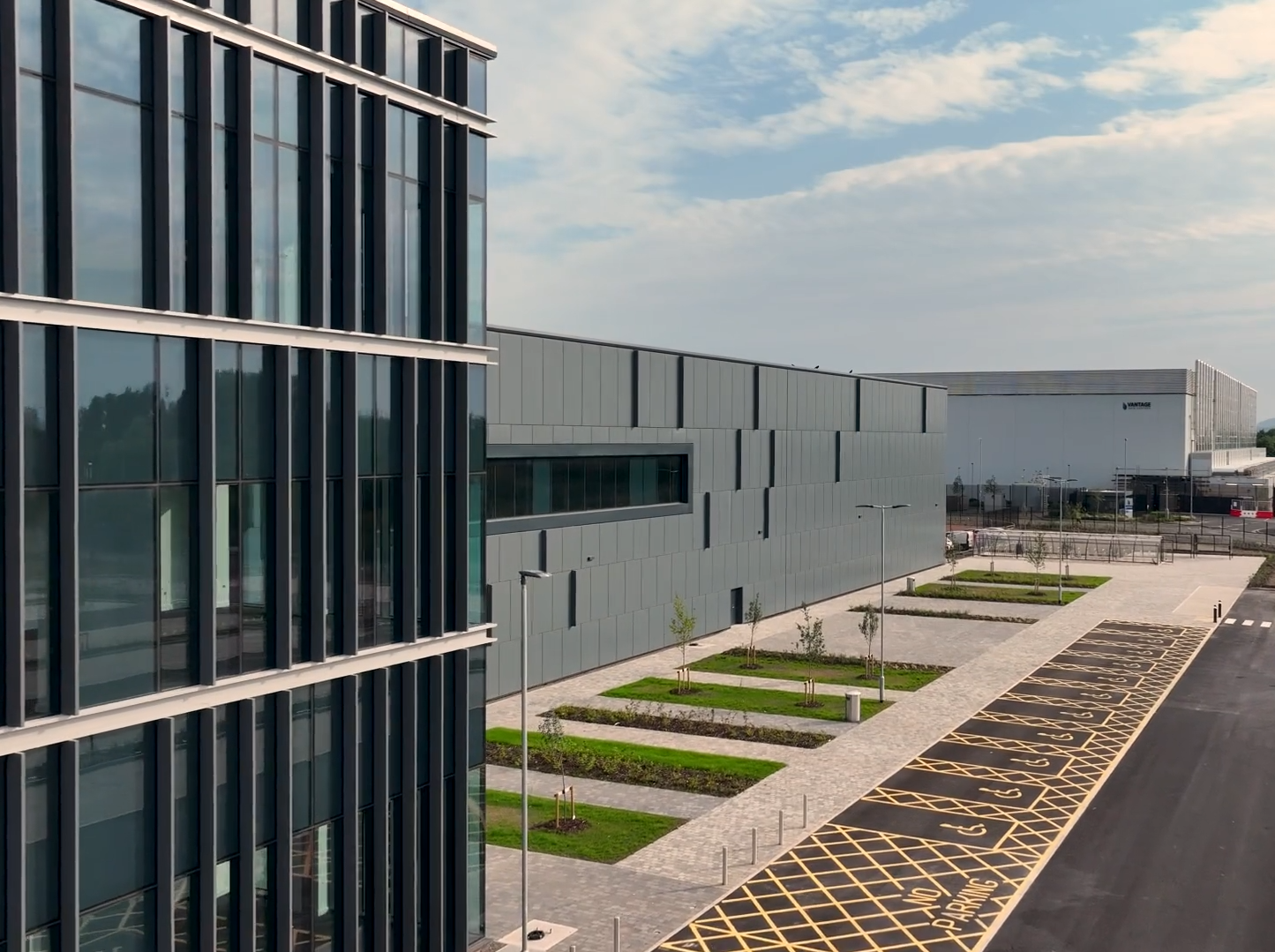 A modern office building with large glass windows stands next to a grey industrial facility. There is a landscaped area with young trees and a parking lot with yellow markings in the foreground under a partly cloudy sky.