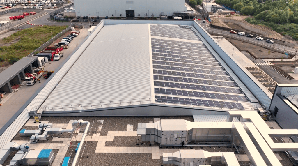 A large industrial building with a sloped roof covered in solar panels, surrounded by parked cars, construction materials, and other industrial structures.