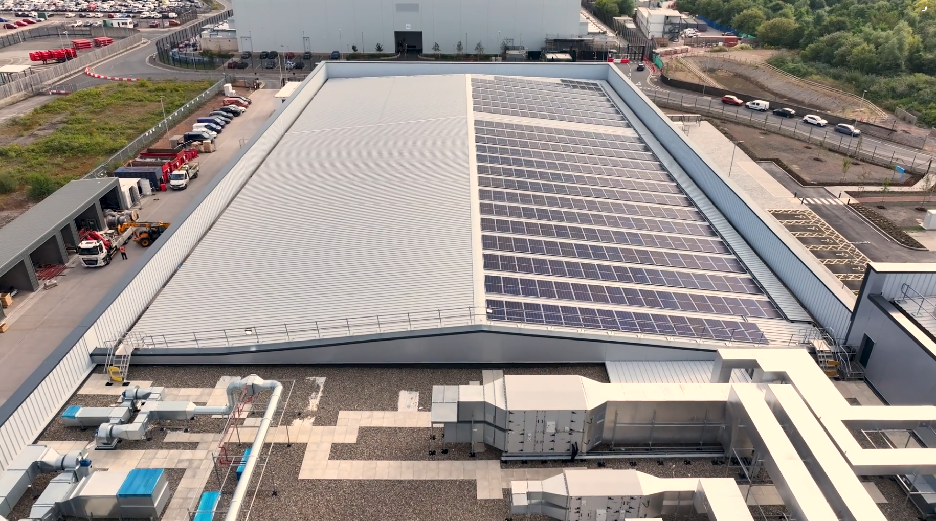 A large industrial building with solar panels installed on one side of its sloped metal roof, surrounded by smaller structures, parked cars, and construction equipment.