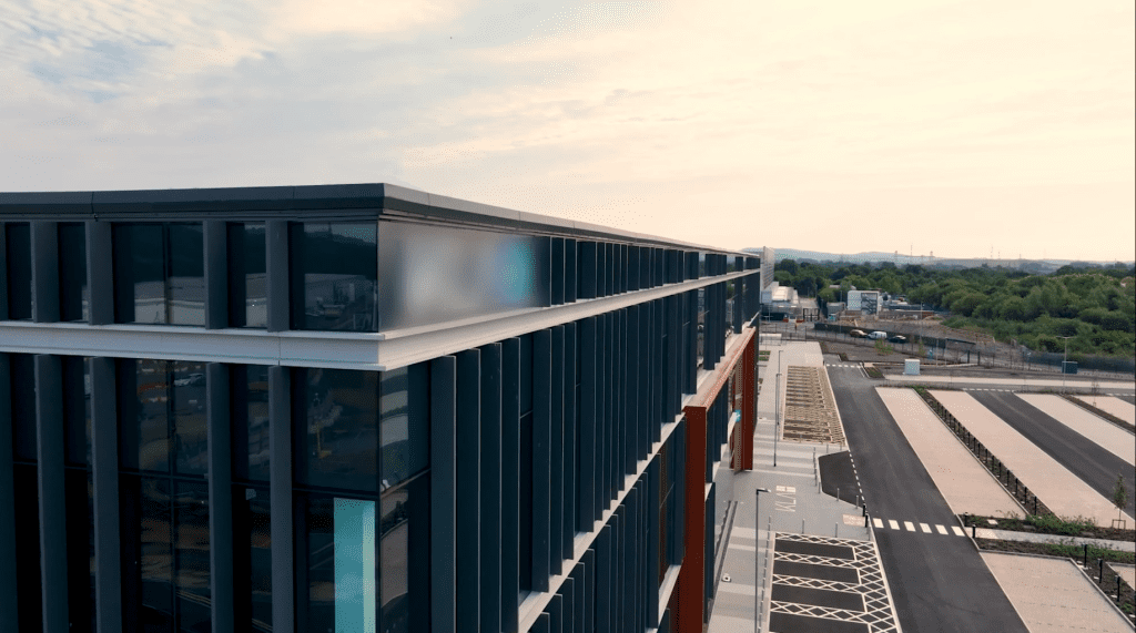 A modern, multi-story building with large glass windows and dark vertical panels, photographed from above near a mostly empty parking lot and a road, with some vehicles and greenery in the distance.
