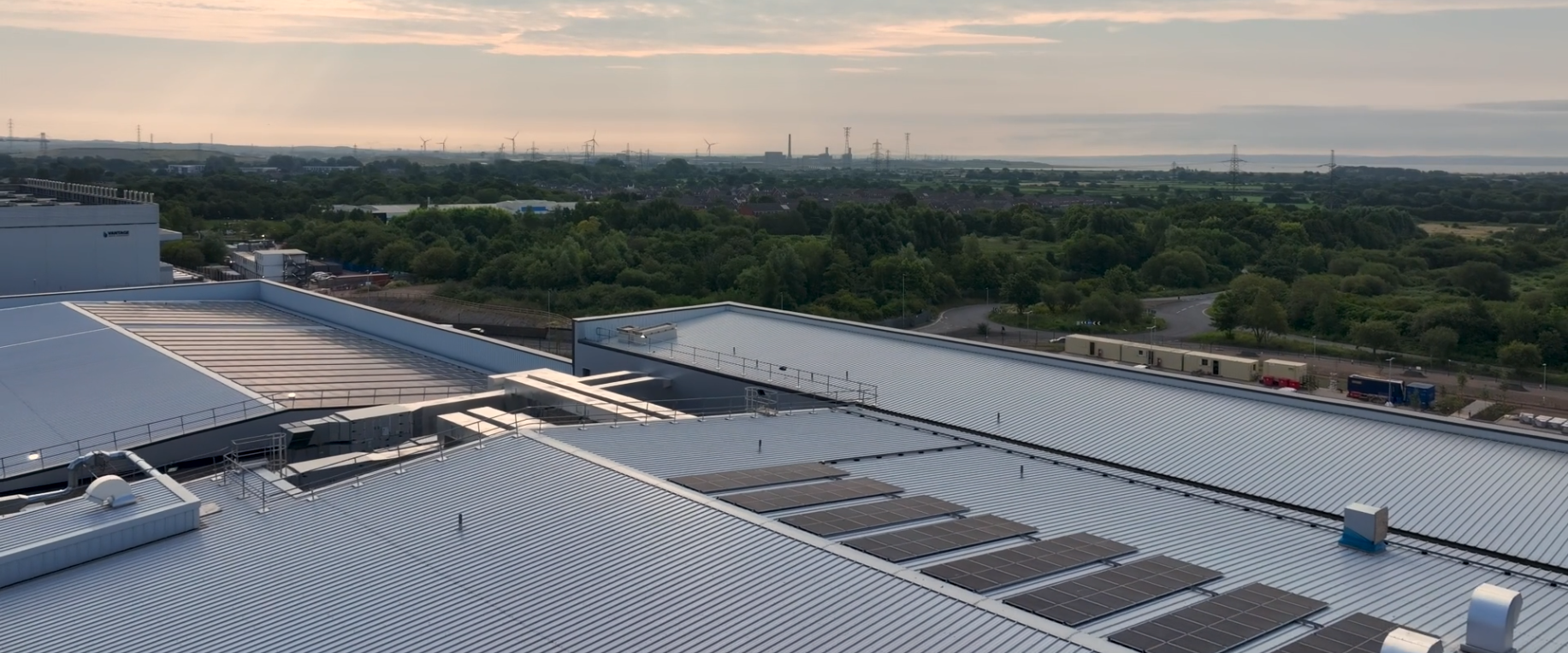 Aerial view of a large industrial building with rows of solar panels on its metal roof, surrounded by greenery and trees, with a distant landscape and wind turbines under a partly cloudy sky at sunset.
