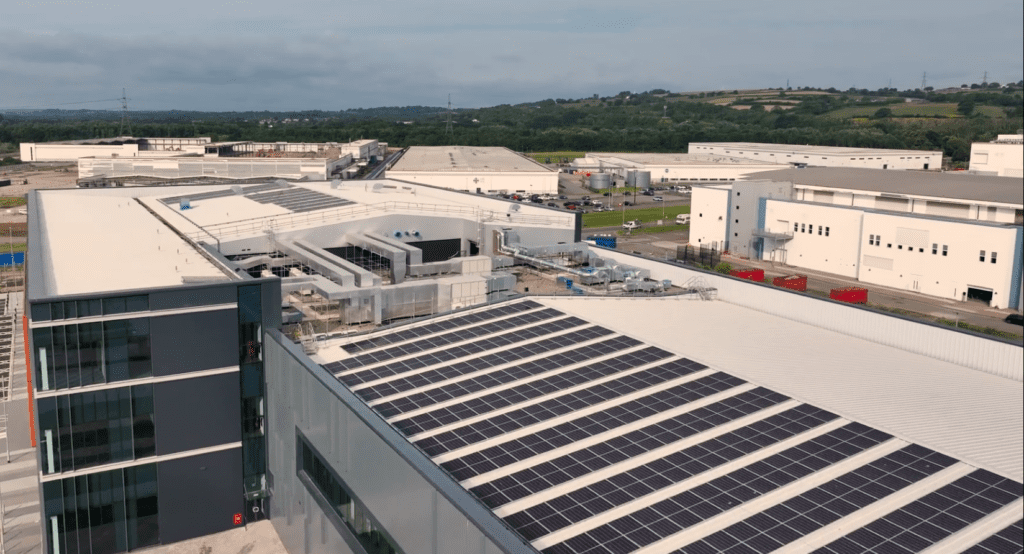 A large industrial complex with white rooftops, some covered in solar panels. Green fields and rolling hills are visible in the background under a cloudy sky.