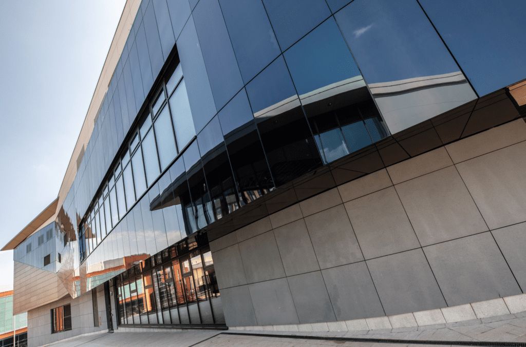 Modern building with large reflective glass panels and gray stone walls, photographed from a low angle on a bright day. The windows reflect surrounding structures and the sky, adding geometric visual interest.