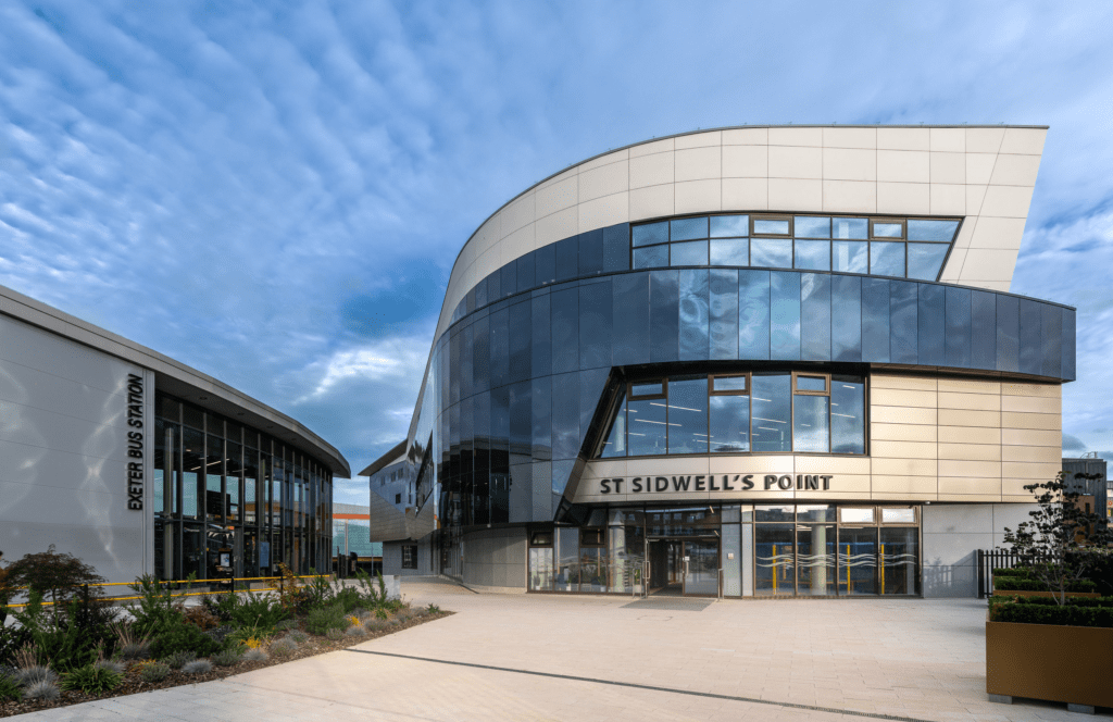 Modern building with curved glass and cream-colored panels labeled "St Sidwell's Point," next to Exeter Bus Station, under a blue sky with scattered clouds. Contemporary landscaping and wide walkway in front.