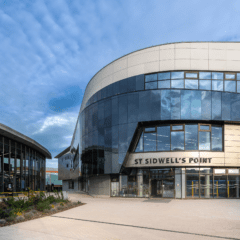 Modern building with curved glass and cream-colored panels labeled "St Sidwell's Point," next to Exeter Bus Station, under a blue sky with scattered clouds. Contemporary landscaping and wide walkway in front.