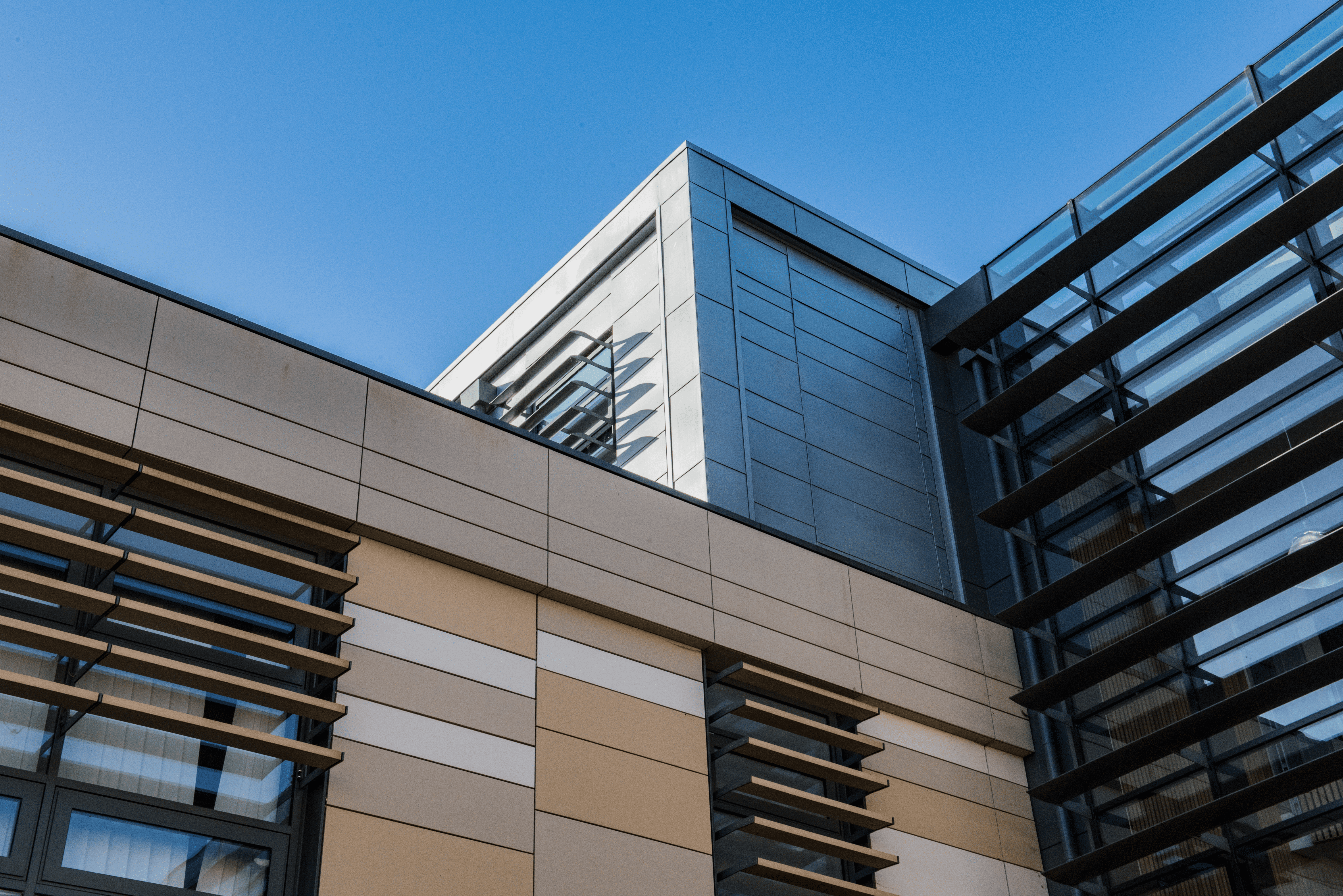 Modern building with beige and gray panels, large windows, and metal sunshades, viewed from below against a clear blue sky. The architecture features angular lines and reflective glass surfaces.