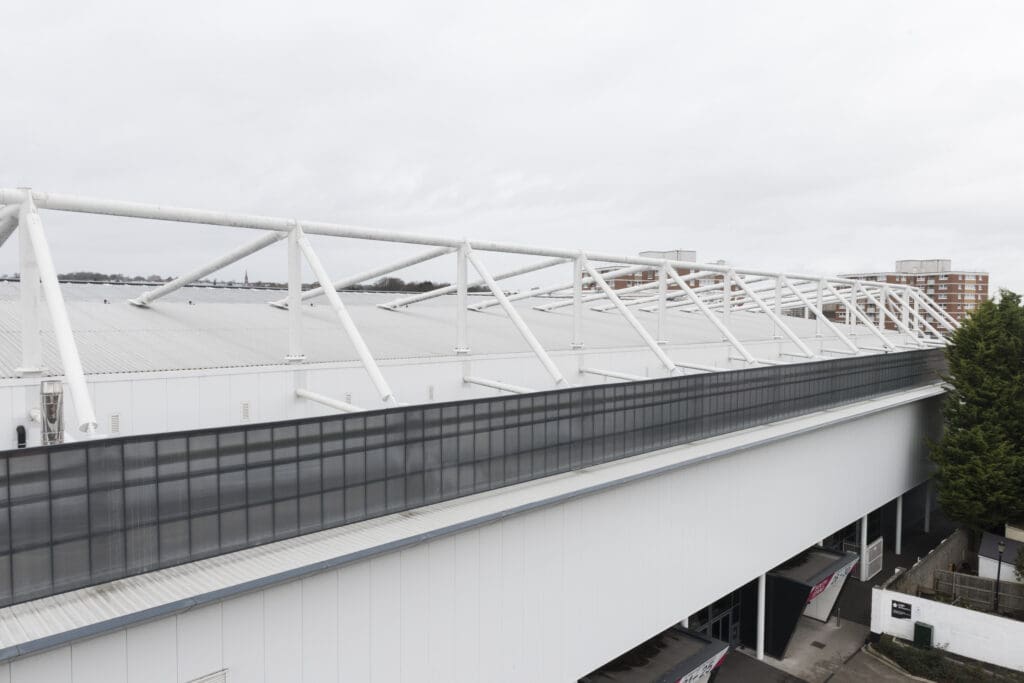 A large white industrial or sports complex building with a modern metal roof structure, tall windows, and surrounding trees, set against an overcast sky.