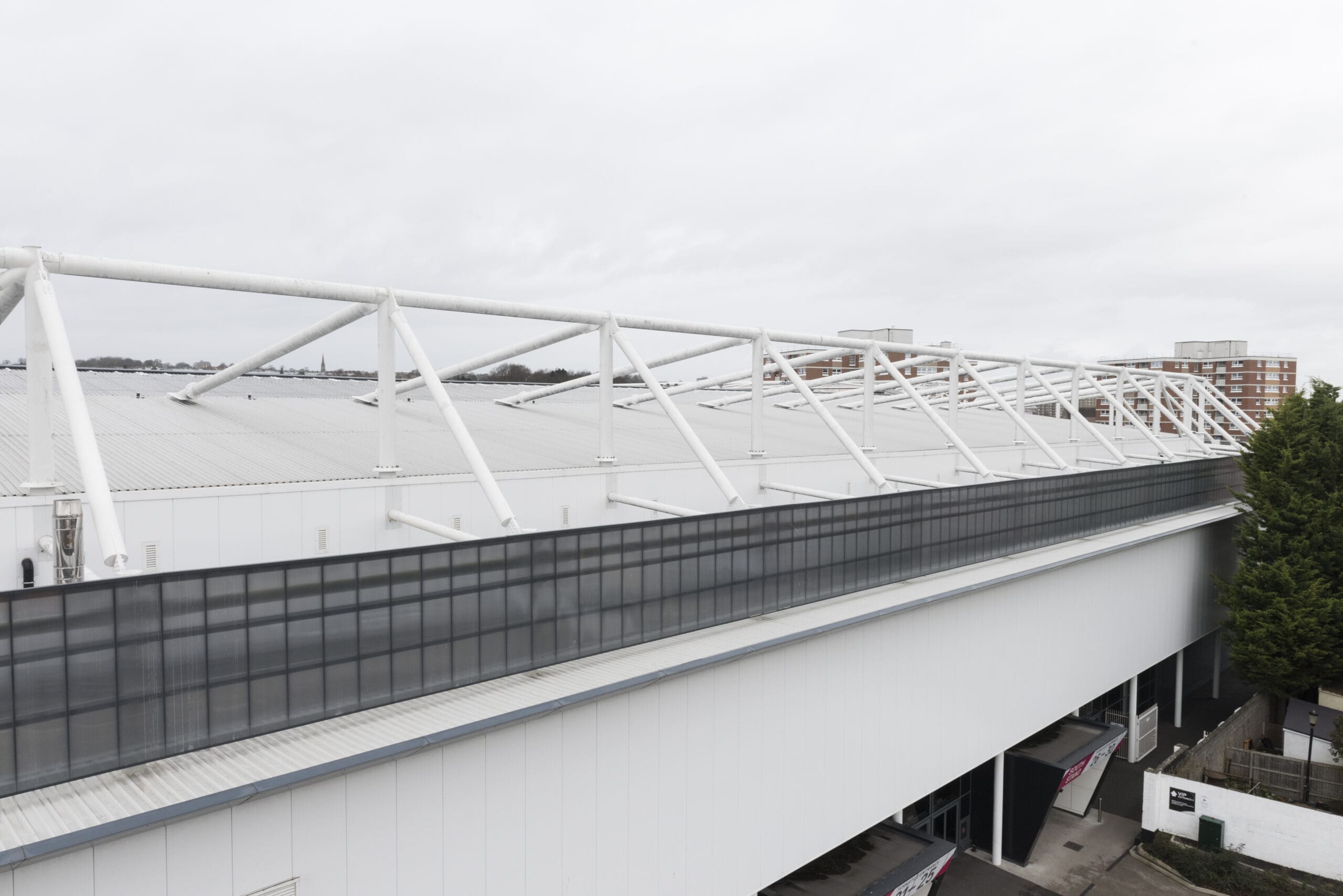 A large white industrial or sports complex building with a modern metal roof structure, tall windows, and surrounding trees, set against an overcast sky.