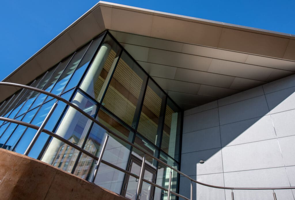 Modern building with large glass windows and a slanted roof, viewed from a low angle against a clear blue sky. A metal railing curves along a concrete ramp in the foreground.