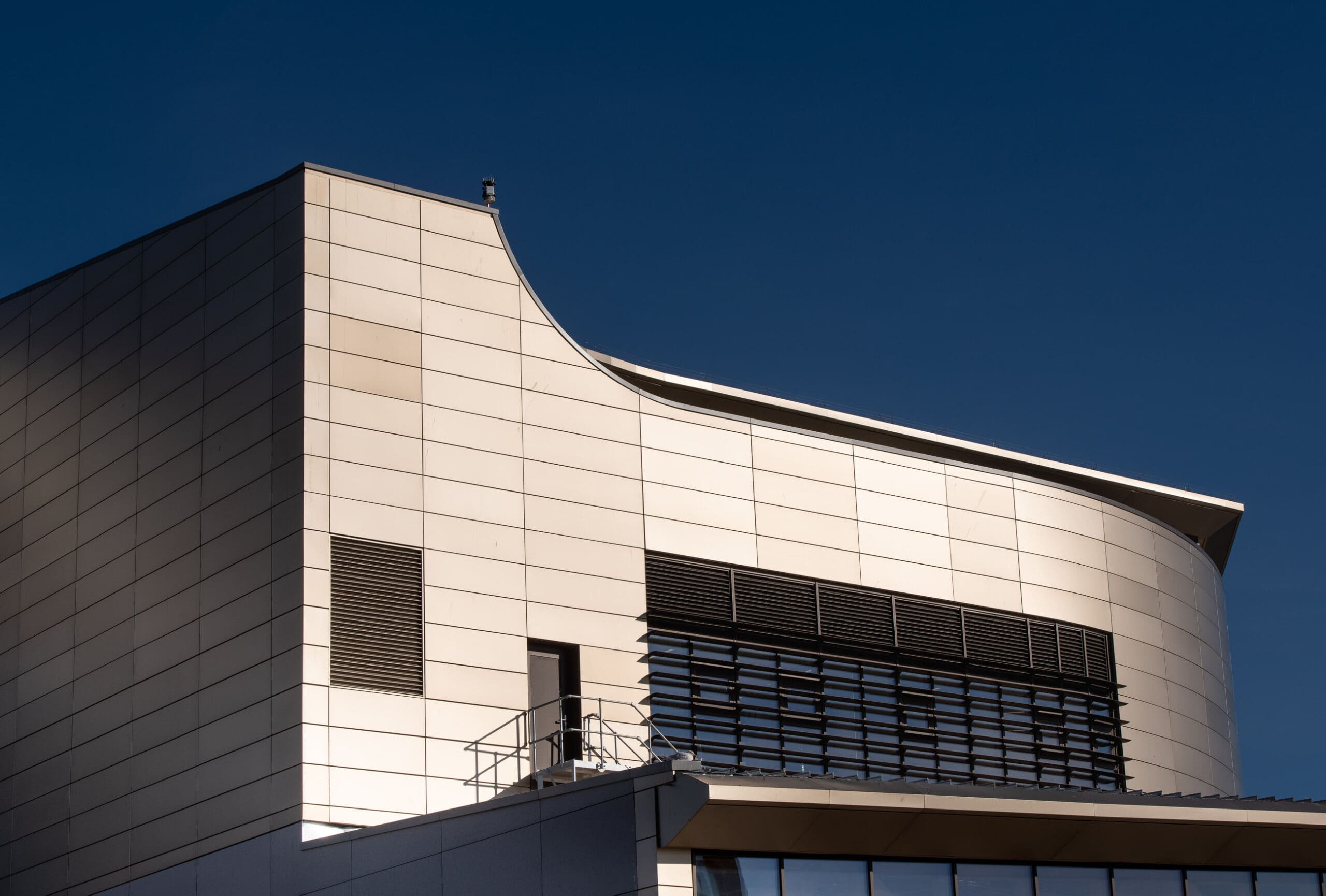 Modern building with curved and angular lines, light-colored exterior panels, black horizontal vent slats, and a small terrace railing, set against a clear deep blue sky.