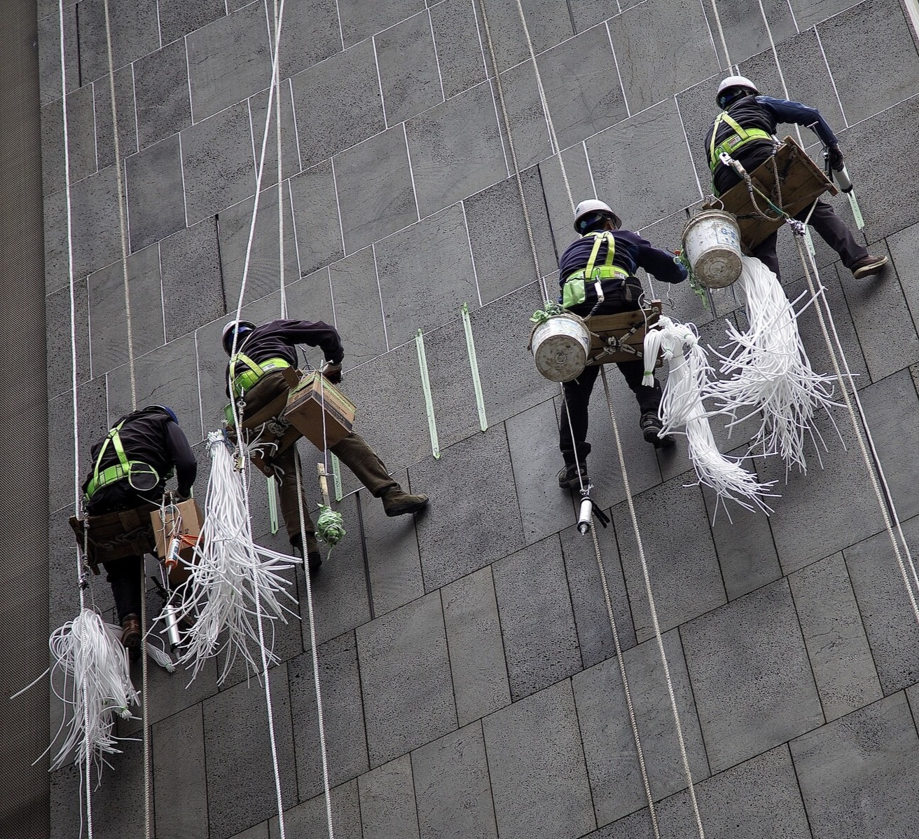 Four workers wearing helmets and harnesses clean or repair a tall tiled building while suspended by ropes, with cleaning tools and buckets hanging beside them. Strips of white material dangle from their equipment.