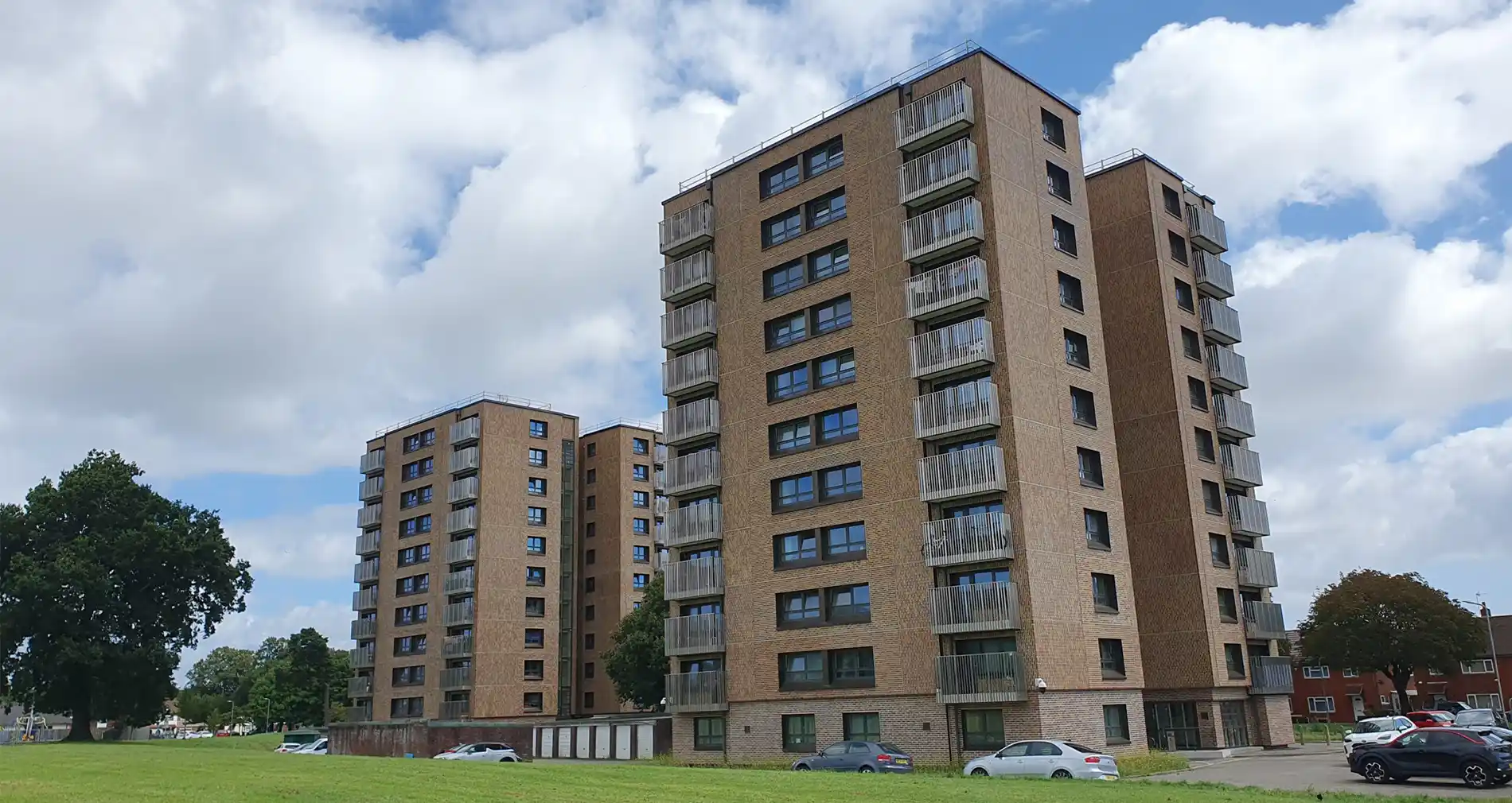 Two mid-rise apartment buildings with brick exteriors and balconies stand side by side on a grassy area, with parked cars and trees nearby under a partly cloudy sky.