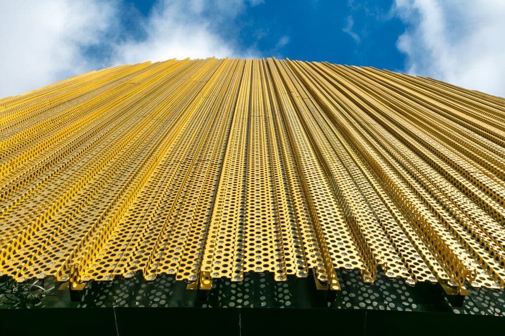 A low-angle view of a modern building facade covered in vertical, perforated gold metal panels against a blue sky with clouds.