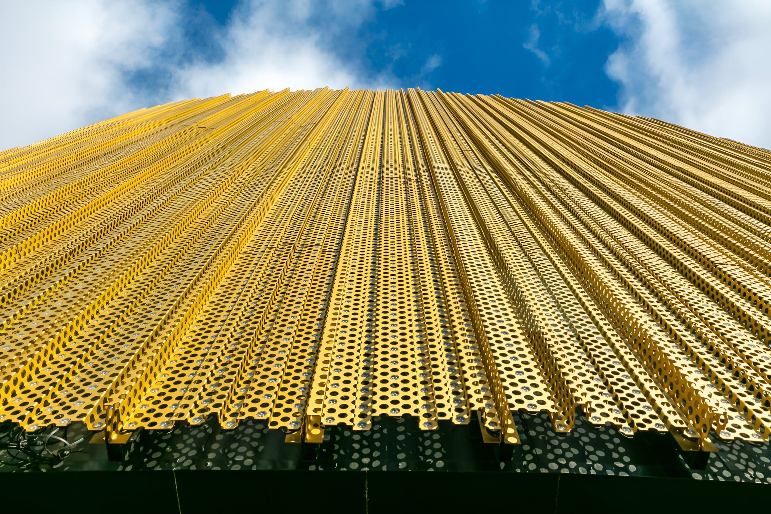 A low-angle view of a modern building facade covered in vertical, perforated gold metal panels against a blue sky with clouds.