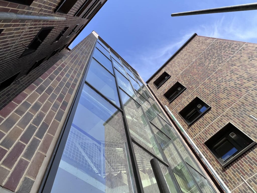 Upward view of three brick buildings with large glass windows and a blue sky above; metal poles are visible in the foreground, adding depth to the architectural perspective.