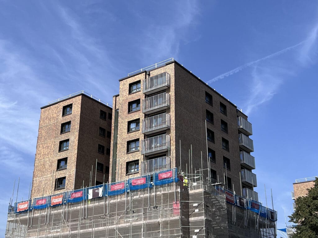 A modern, mid-rise brick apartment building under construction, surrounded by scaffolding and safety netting, set against a clear blue sky.