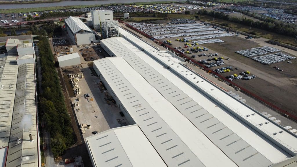 Aerial view of a large industrial facility with long white-roofed buildings, outdoor storage areas, parked vehicles, and a river in the background. Trees border one side of the complex.
