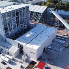Aerial view of an industrial facility with large white and gray buildings, steel frameworks, pipes, construction materials, and vehicles on the grounds; conveyor belt structure cuts across the site. Trees and parked cars are visible in the background.