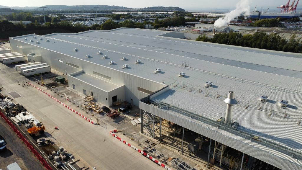 A large industrial factory with a metal roof, surrounded by construction equipment and vehicles. Smokestacks emit steam in the background, and trees border the site. The area appears to be under active development.