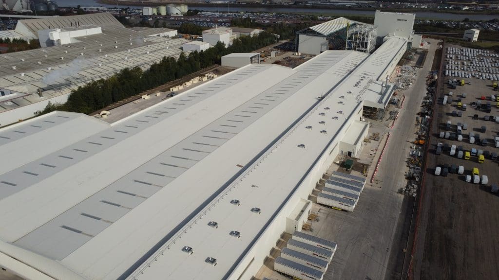 Aerial view of a large industrial warehouse complex with long white roofs, loading docks, parked trucks, and adjacent parked vehicles; trees and other industrial buildings are visible in the background.
