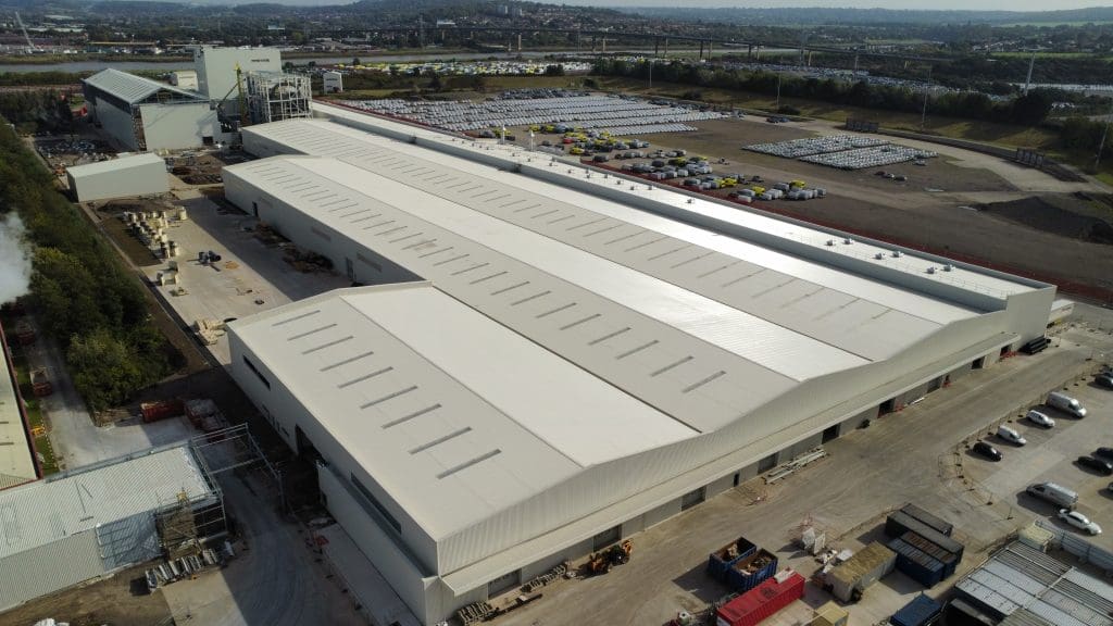 Aerial view of a large industrial warehouse with a white roof, surrounded by parked cars and adjacent to open land and smaller buildings, with a cityscape and roads visible in the background.