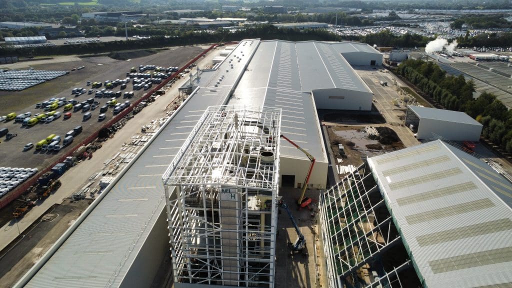 Aerial view of a large industrial complex with warehouses, a steel-framed structure under construction, parked vehicles, and green fields in the background.