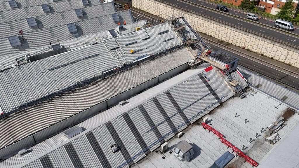 Aerial view of a building with a partially collapsed metal roof, construction scaffolding, and debris. Nearby, there is a road with moving vehicles and a sound barrier wall.
