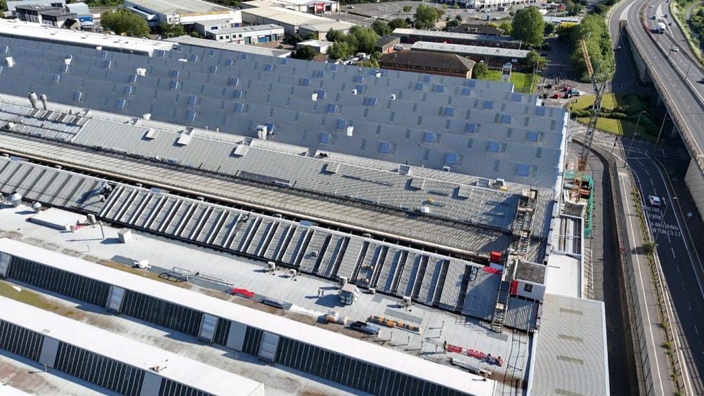 Aerial view of a large industrial building with a slanted roof and multiple skylights, adjacent to railway tracks. Cars and roads are visible on the right, with additional buildings and trees in the background.