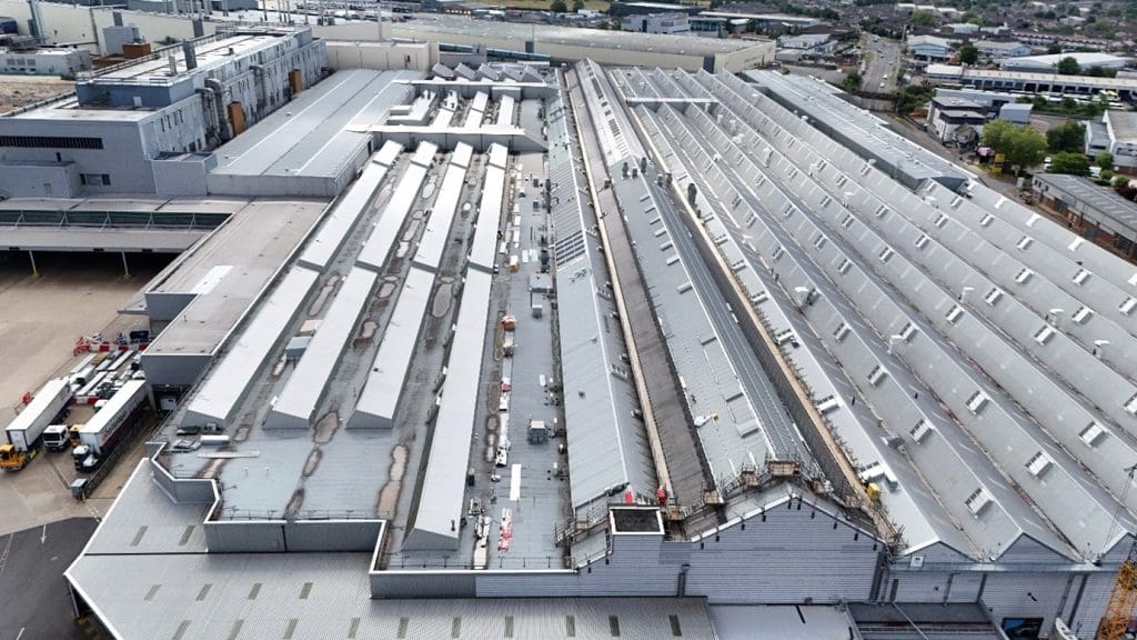 Aerial view of a large industrial complex with multiple long, grey factory buildings featuring sawtooth roofs and skylights, surrounded by service vehicles and adjacent to other industrial structures.