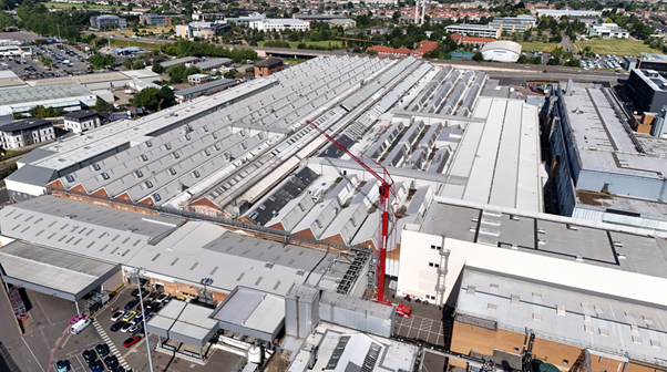 A large industrial complex with rows of rooftops, skylights, and a red construction crane extending over the buildings, surrounded by parking lots and greenery in the background.