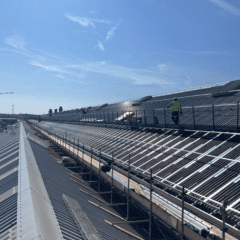 Workers in high-visibility vests install panels or carry out maintenance on a large industrial metal roof under a clear blue sky, with scaffolding and safety railings visible along the roof edge.