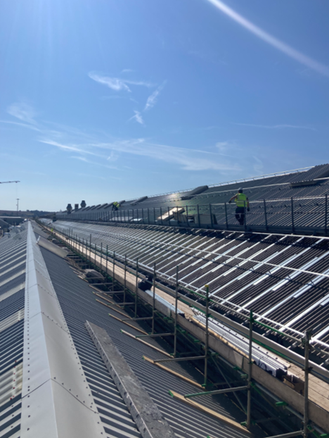 Workers in high-visibility vests install panels or carry out maintenance on a large industrial metal roof under a clear blue sky, with scaffolding and safety railings visible along the roof edge.