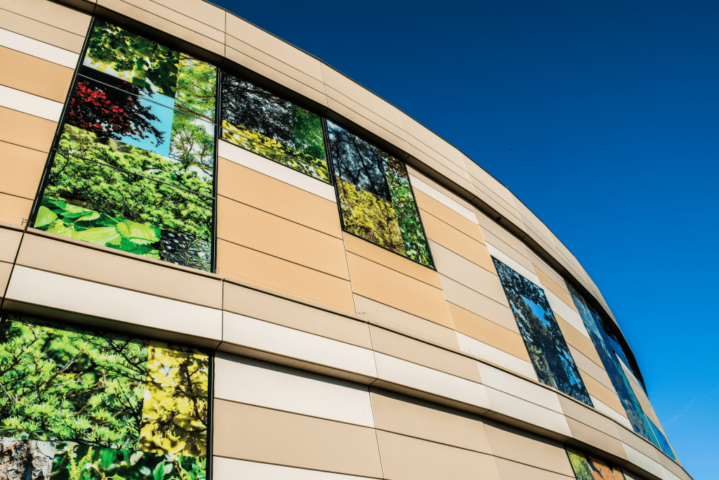 A modern beige building with vertical windows displaying vibrant nature scenes, including green foliage and trees, set against a clear blue sky.
