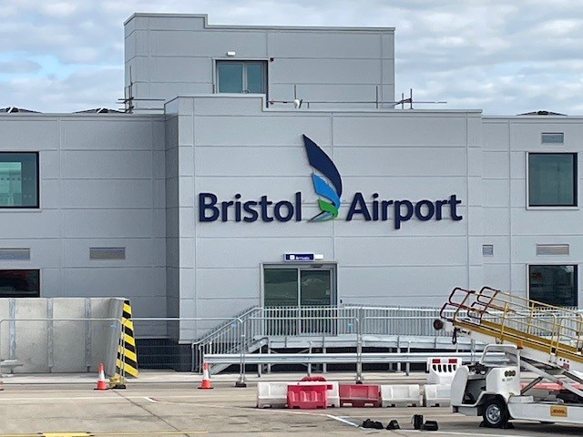 A view of the exterior of Bristol Airport, showing the main building with "Bristol Airport" signage and a blue and green logo, with airport ground equipment and barriers in the foreground.