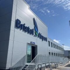 Exterior of Bristol Airport building with large blue sign, glass doors, and metal stairs under a sunny sky with some clouds.