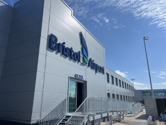 Exterior of Bristol Airport building with large blue sign, glass doors, and metal stairs under a sunny sky with some clouds.