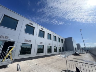 A modern, gray, two-story building with large windows under a blue sky with wispy clouds. Metal barricades and a staircase are visible near the entrance, with concrete pavement in the foreground.
