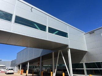 A modern, silver industrial building with large windows and an elevated walkway under a clear blue sky. Yellow bollards are placed near the entrance.