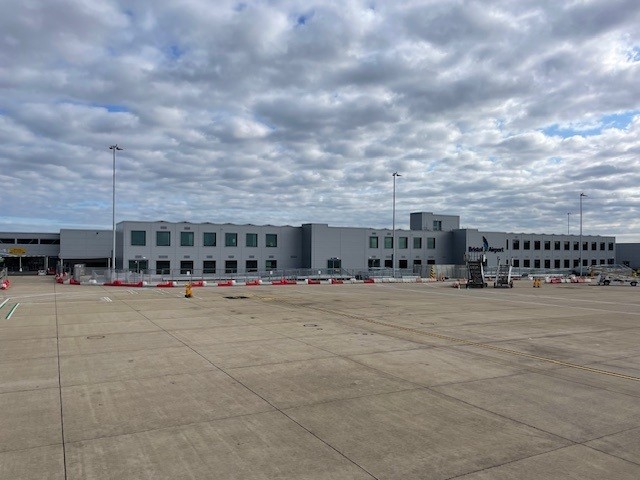 A large, modern airport terminal with a gray facade sits under a cloudy sky, viewed from the tarmac. The foreground shows an empty concrete apron with some construction barriers and equipment.