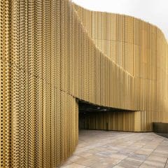 Curved modern building with a gold perforated metal facade, creating an intricate pattern. The walkway in front is paved, and the sky is cloudy, hinting at an urban setting in the background.