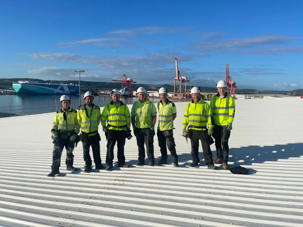 Six workers in high-visibility jackets and helmets stand on a large corrugated metal roof, with cranes, a ship, and blue sky in the background.